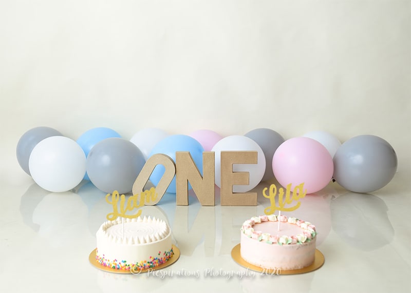 a photo celebrating the first birthday of two babies, there are two cakes in the foreground, balloons in the back, with cardboard letters spelling ONE